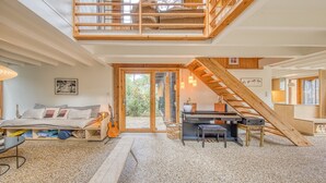 Ein gemütliches Interieur mit Schlafzimmer im Zwischengeschoss, einem Klavier, einer Treppe und Blick durch eine Glastür.