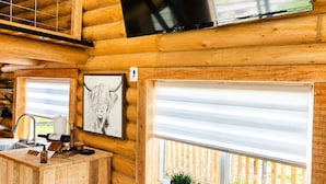 A log cabin interior with a wooden bar, a television mounted on the wall, and a window with a view of a wooden deck.