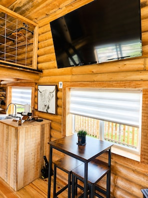 Ein rustikales Blockhaus-Interieur mit einer Holztheke, einem an der Wand befestigten Fernseher und einem Fenster mit Blick auf eine Holzterrasse.