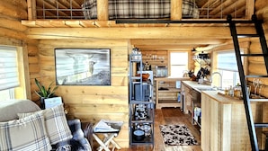 A cozy cabin interior with a loft bed, a dining area, a kitchen, and a living room with a sofa and a cowhide rug.