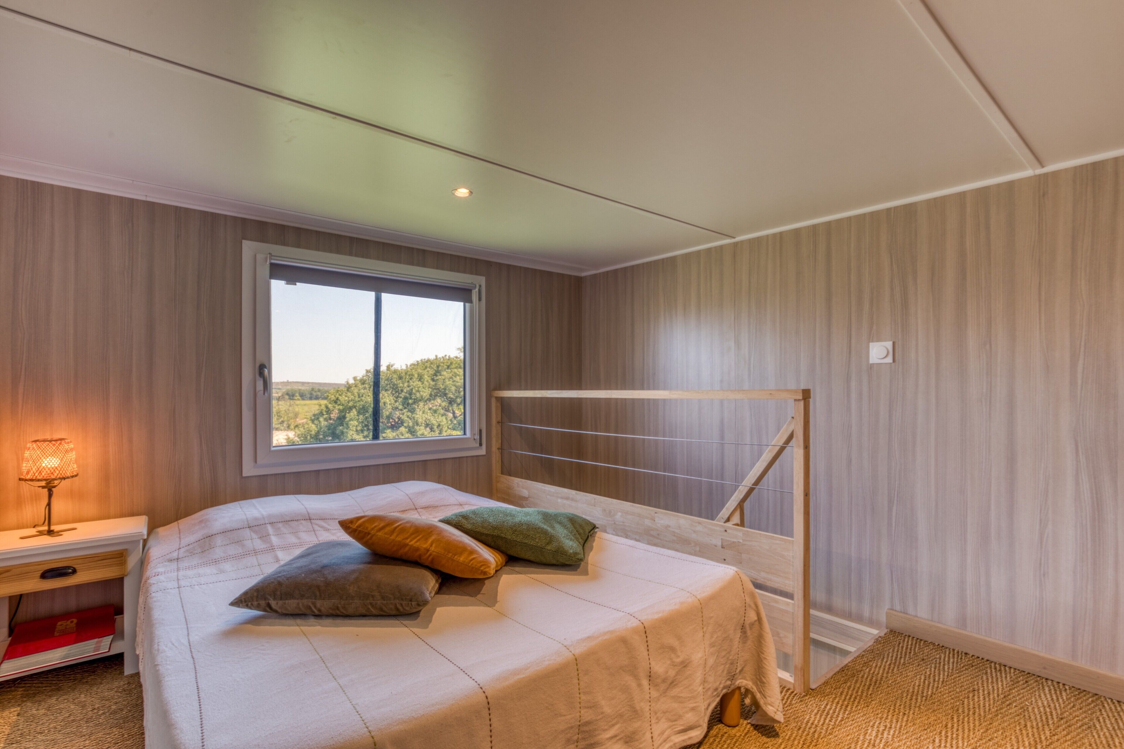 Ein Schlafzimmer mit Bett, Kissen, einem Nachttisch, einem Fenster und Blick ins Grüne.