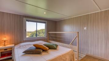 Ein Schlafzimmer mit Bett, Kissen, einem Nachttisch, einem Fenster und Blick ins Grüne.
