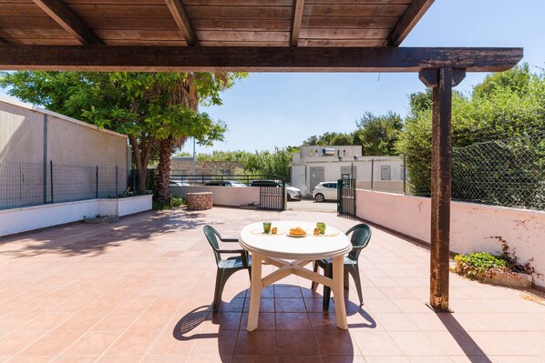 An outdoor patio with a round table and chairs, a pergola overhead, and a view of the surrounding area including a fence and trees.