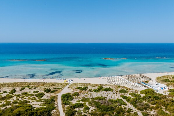An aerial view of a beach with a clear sky, a sandy shore, and a building with a blue roof.
