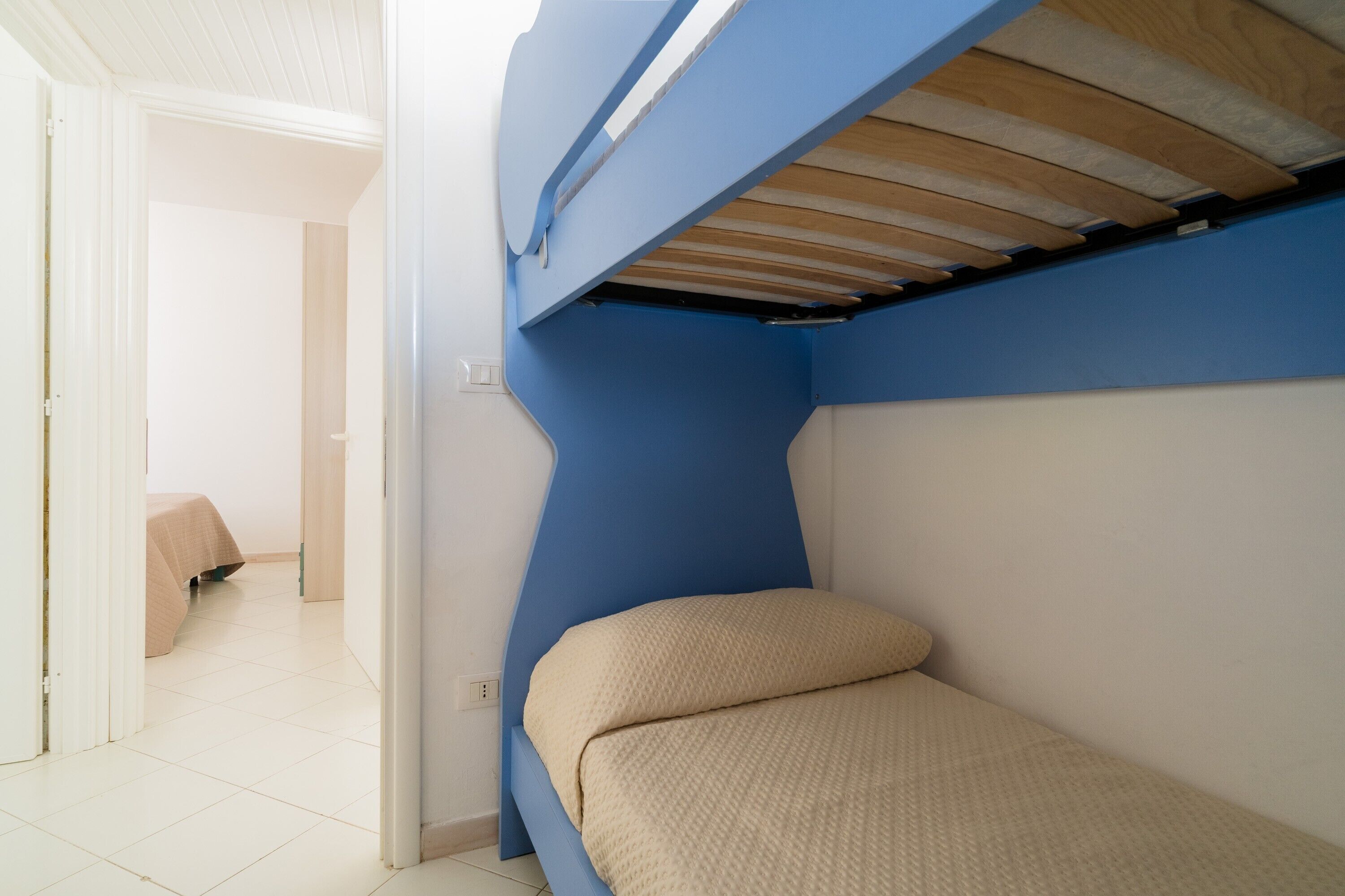 Ein Etagenbett mit blauer Kopfteil und einer beigen Matratze, eine weiße Tür und ein gefliester Boden.