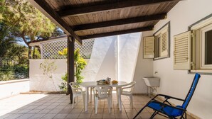 Un patio techado con una mesa redonda y sillas, una silla plegable azul, un lavamanos y una ventana con contraventanas.