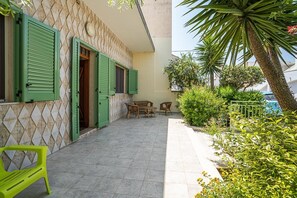 Un bâtiment avec des volets verts, une terrasse carrelée, des meubles en osier et un palmier.