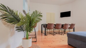 Uma sala de estar moderna com um sofá cinzento, uma área de jantar com cadeiras castanhas, uma televisão fixada na parede e uma grande planta em vaso.