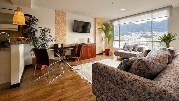 Un salon moderne avec un canapé, une table à manger et des chaises. Un téléviseur est fixé au mur, il y a une fenêtre avec vue sur les montagnes, et un coin cuisine avec un évier et des placards.