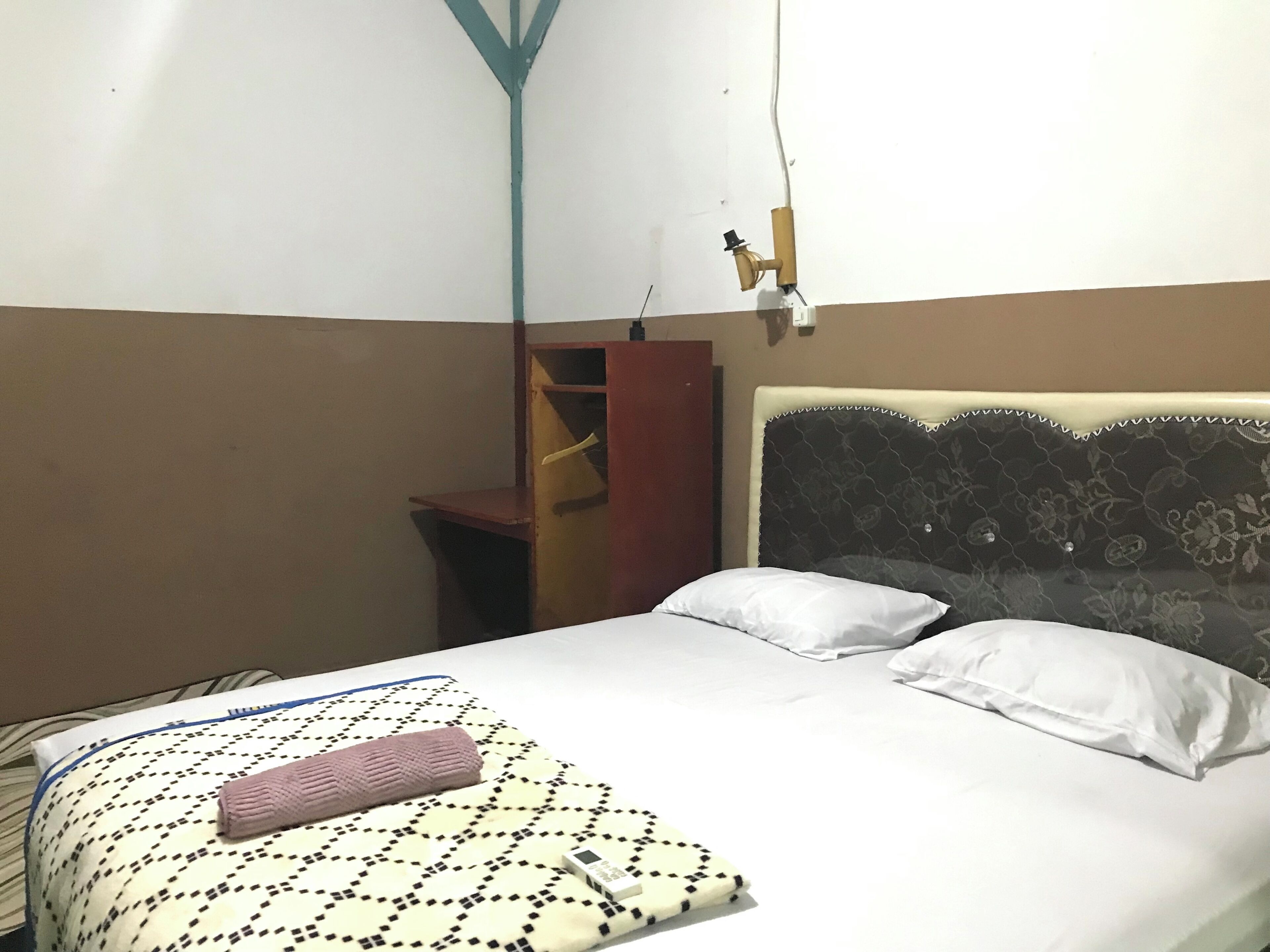 Un lit simple avec une couvre-lit à motifs, deux oreillers blancs, une tête de lit ornée de motifs floraux et une petite étagère en bois.