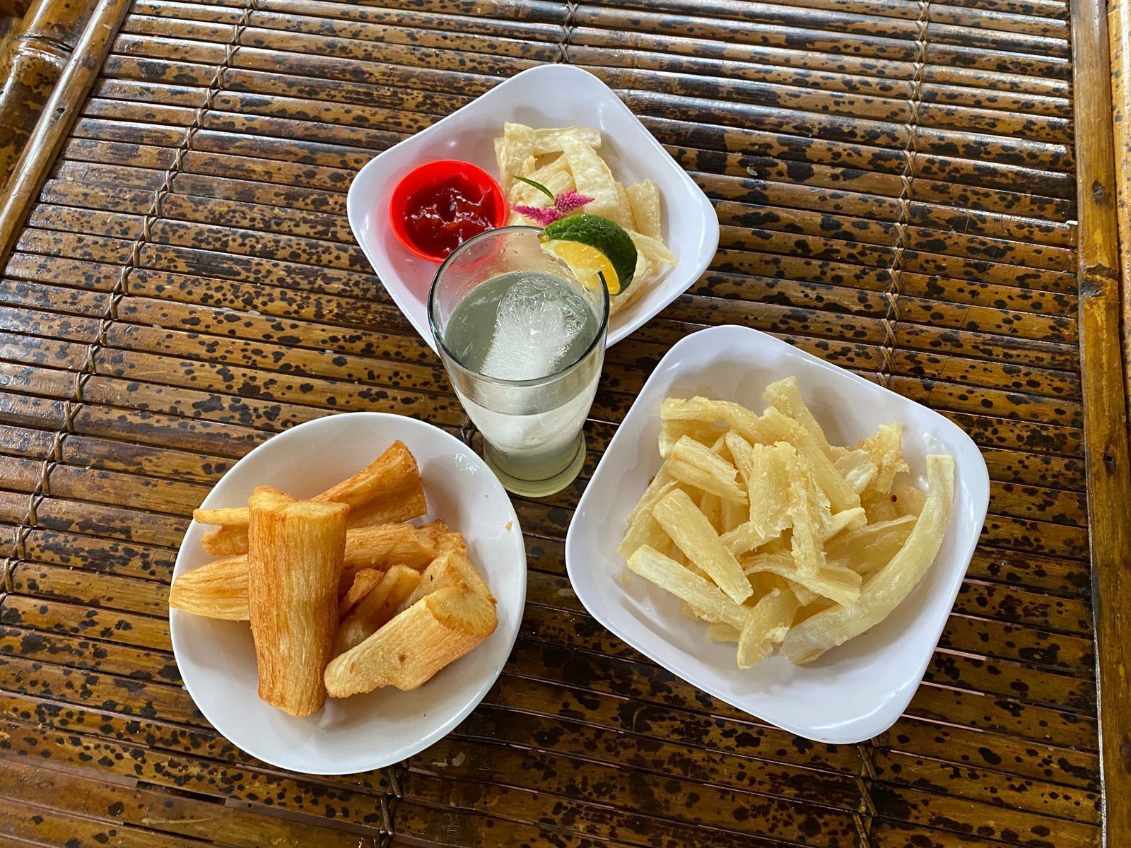 Three white bowls containing different food items, a glass of water, and a red sauce on a bamboo mat.