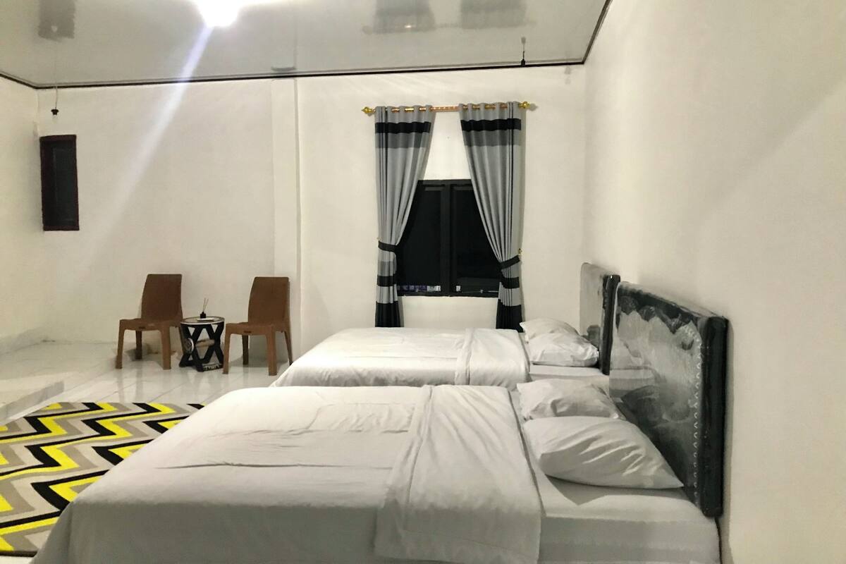 A hotel room with two beds, a small table with chairs, a window with curtains, and a painting on the wall.