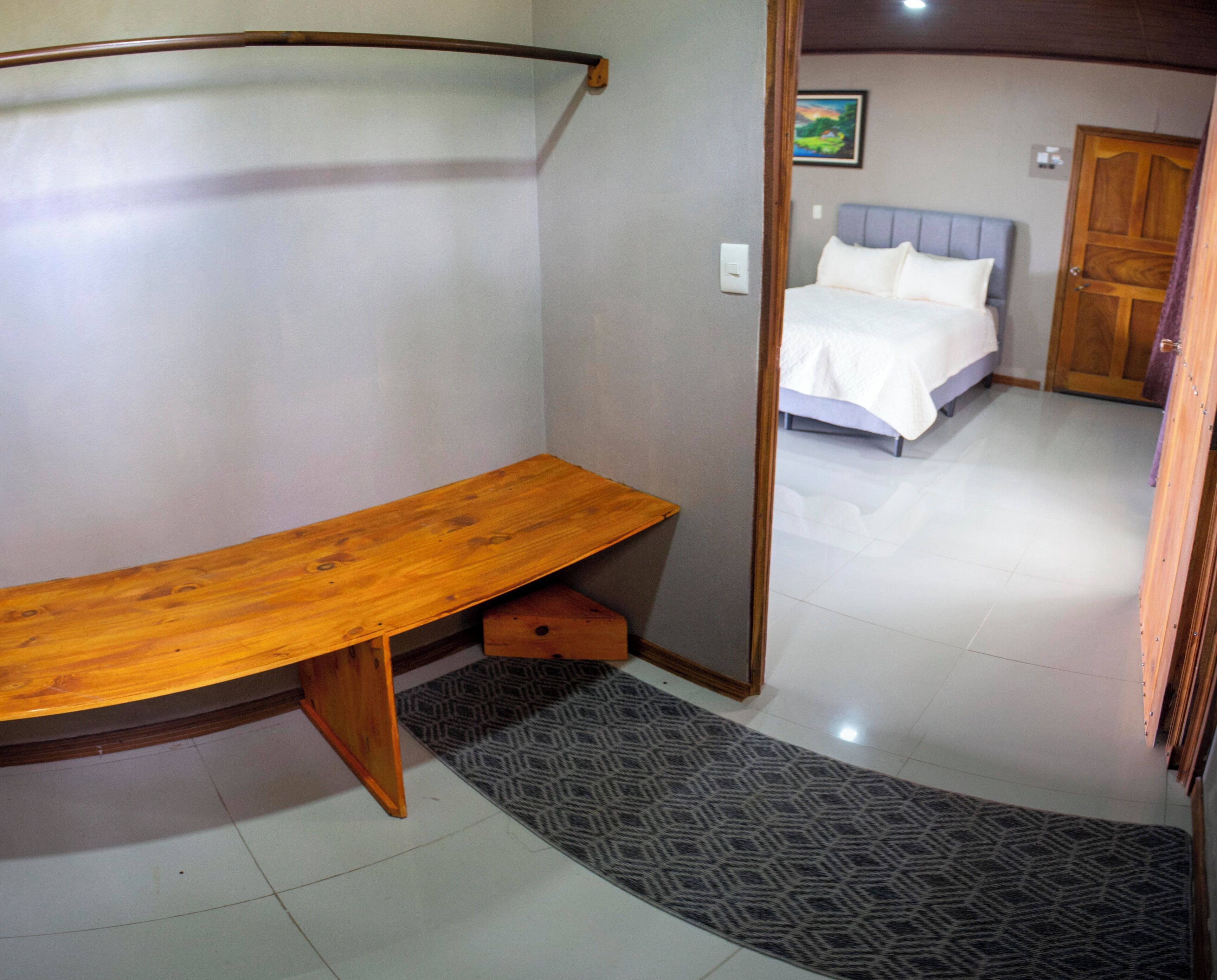 Quarto com um banco de madeira, tapete estampado, cama com roupa de cama branca e porta que dá para outro cômodo.
