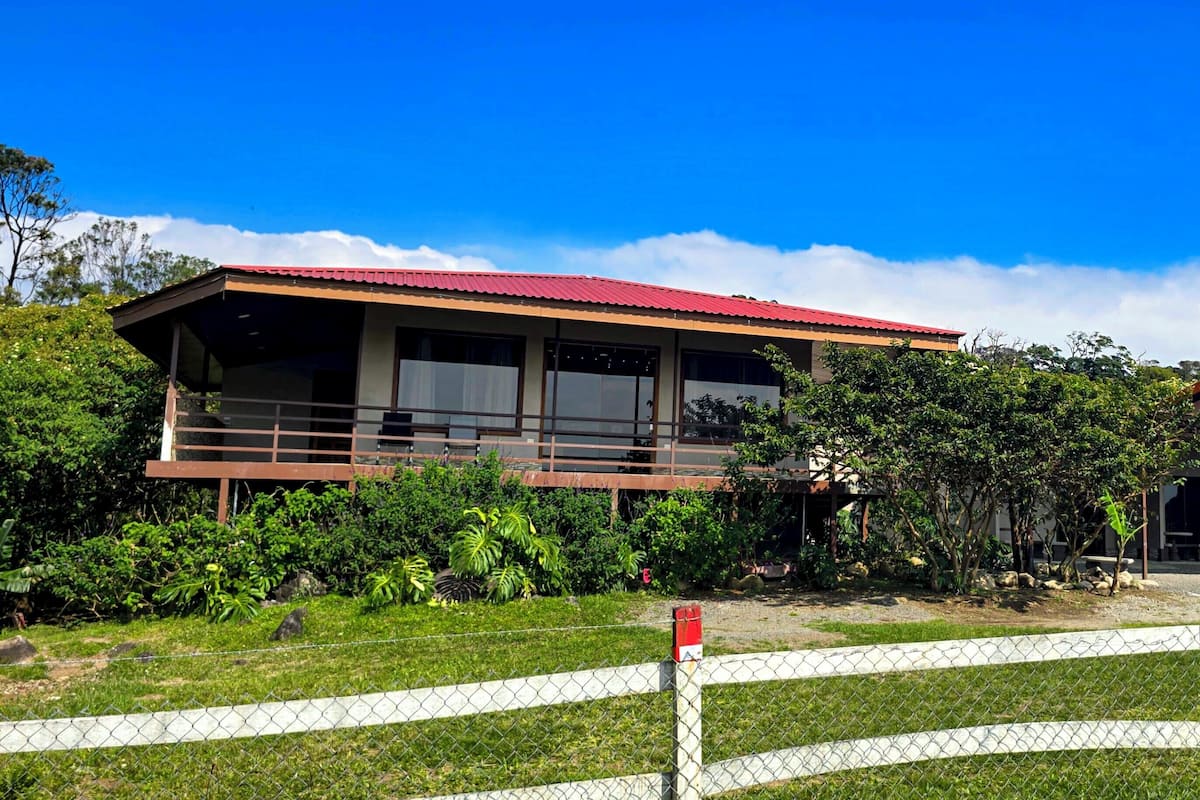 A two-story building with a red roof, surrounded by lush greenery and a white fence.
