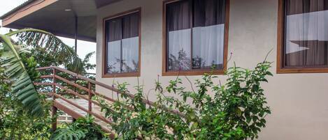 A house with a covered porch, a stone pathway, and lush greenery including banana plants and ferns.