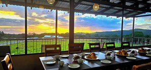 A dining area with a table set for breakfast, overlooking a lush green landscape and hills during sunset. - Almore Hotel (Monteverde)