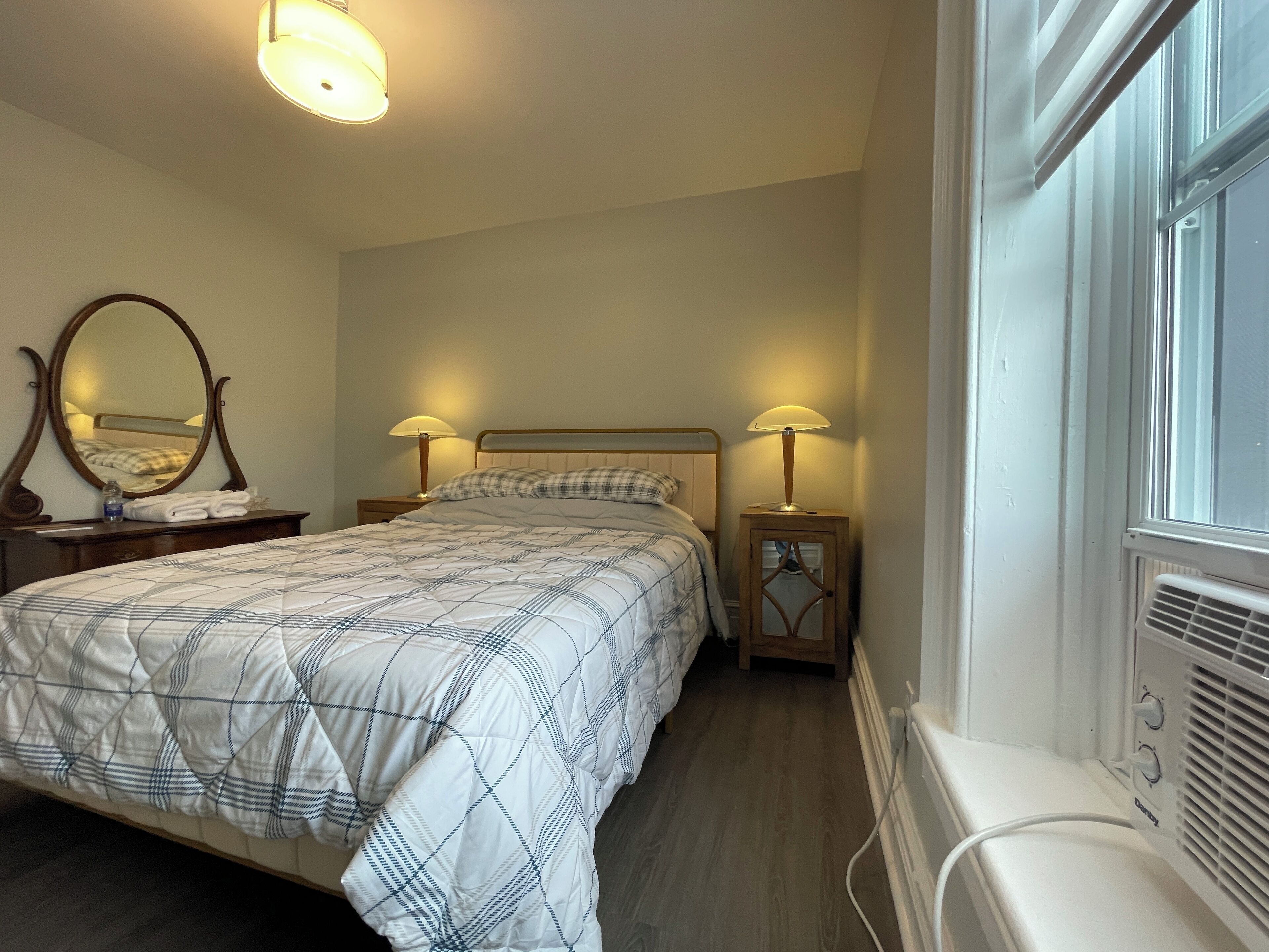 A neatly made bed with a checkered duvet, two bedside lamps, a mirror, and a radiator.