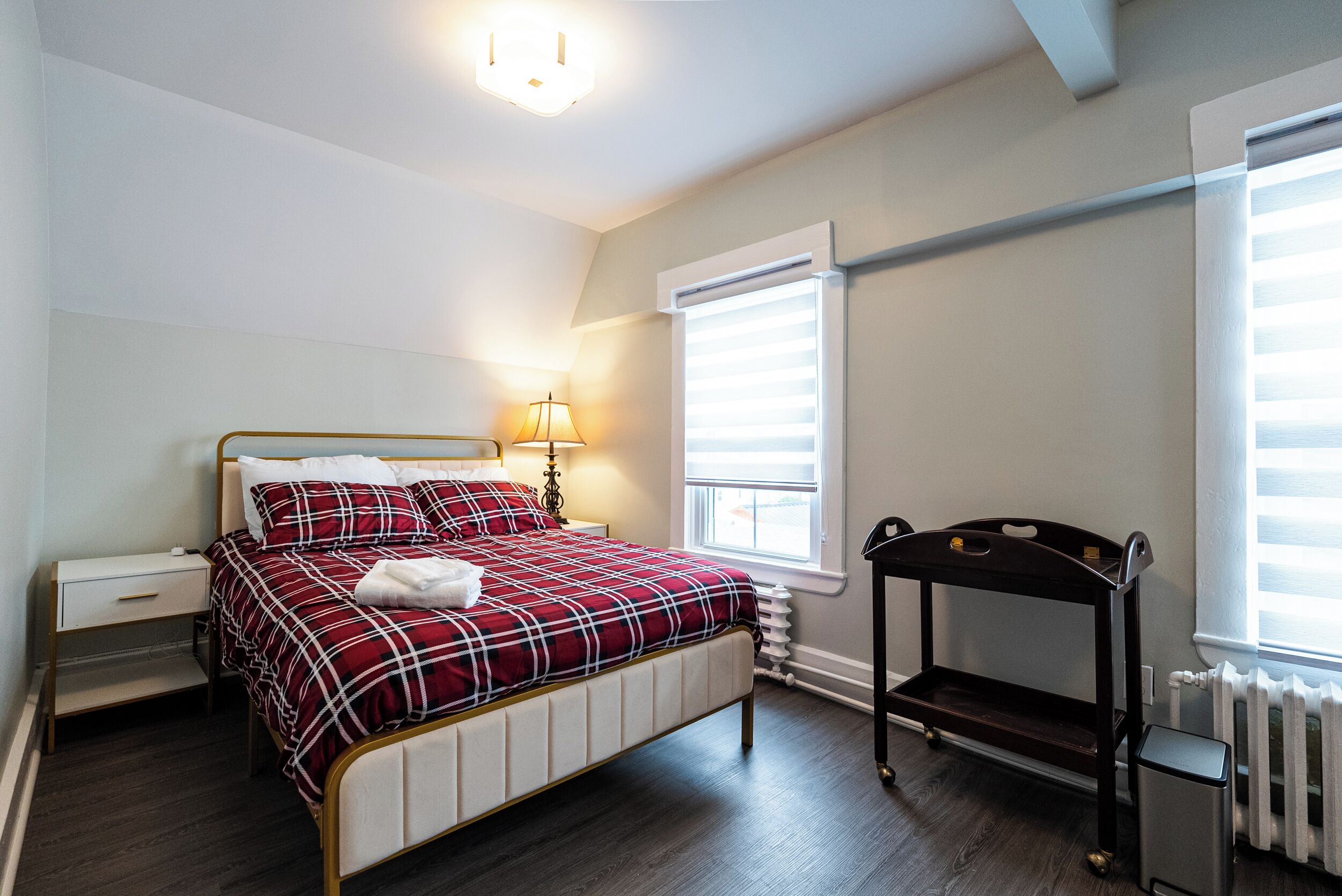 Une chambre à coucher comprenant un lit, une table de chevet, une lampe, un radiateur et des stores aux fenêtres.