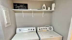A laundry room with two white front-loading washers and dryers, a shelf above with a 'Together' sign, and a white door on the left.