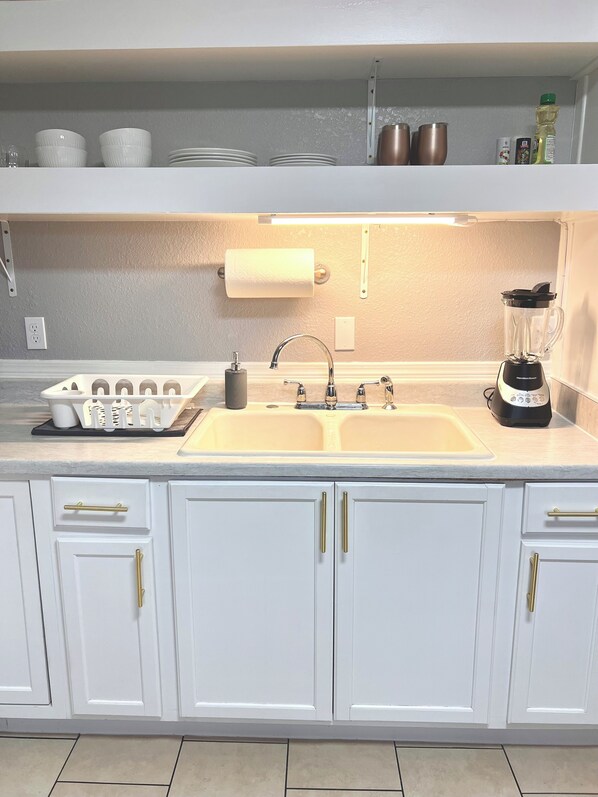 A kitchen with white cabinets, a double sink, a blender, and a shelf with dishes and cookware.