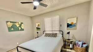 A bedroom with a bed, a ceiling fan, a window with blinds, a bedside table with a lamp, a wall map, and a decorative pillow.