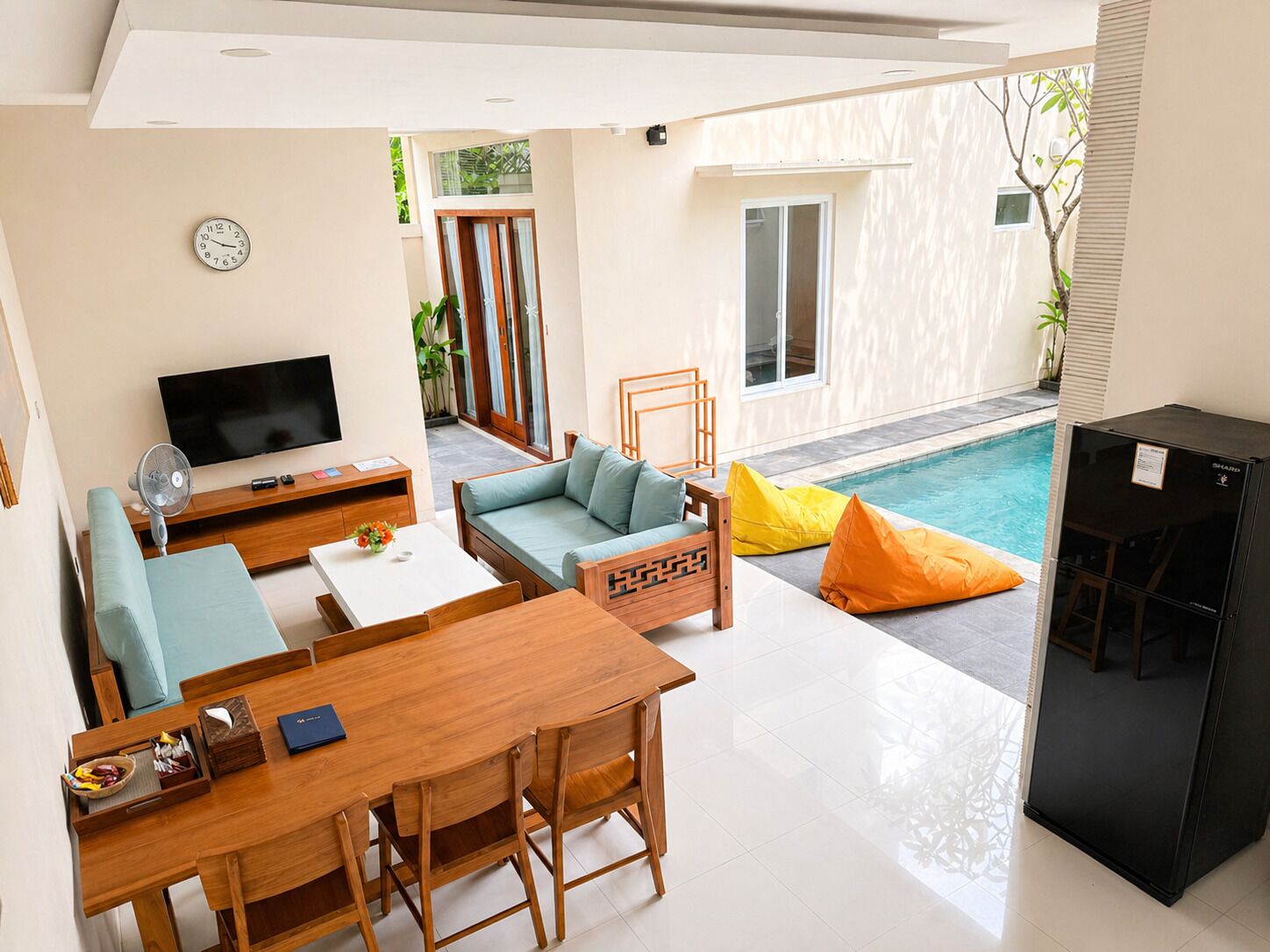 Een moderne woonkamer met een televisie, bank, eettafel en stoelen. Achtergrond is een zwembad.