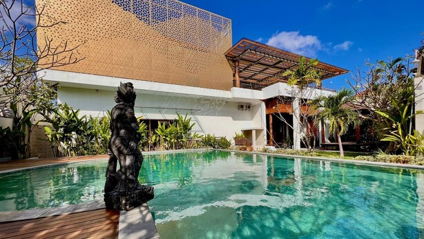 Piscina com uma estátua, cercada por plantas tropicais e um edifício moderno com fachada geométrica.