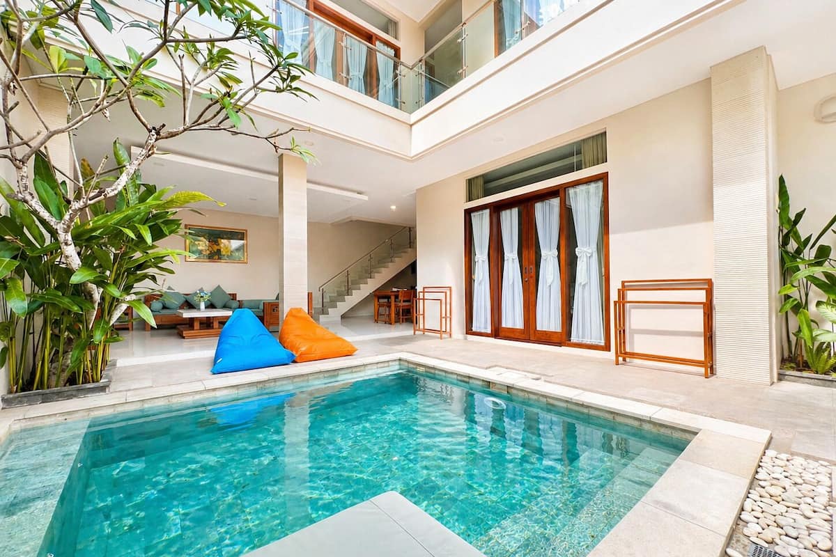Une piscine intérieure moderne, dotée d’une eau cristalline, entourée d’un espace détente avec des coussins, un escalier et de grandes fenêtres.