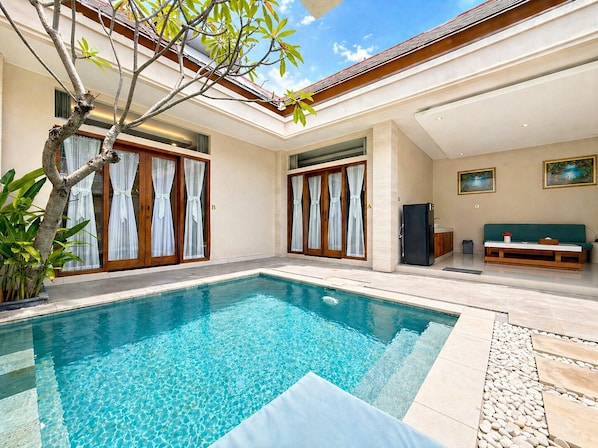 Una piscina con cubierta de baldosas, un sofá verde y un refrigerador en una habitación moderna con ventanas amplias y cortinas.