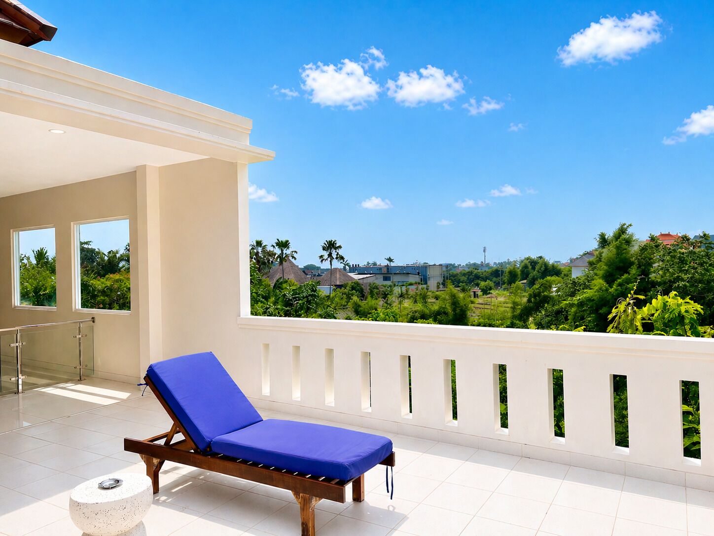 A balcony with a blue lounge chair, a white balustrade, and a view of a green landscape with buildings in the distance.
