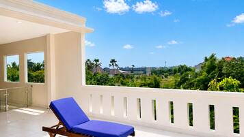 A balcony with a blue lounge chair, a white balustrade, and a view of a green landscape with buildings in the distance.
