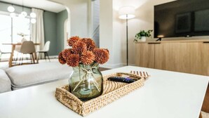 Ein modernes Wohnzimmer mit einem weißen Sofa, einer Glaskaraffe mit Trockenblumen, einer Fernbedienung und einer gewebten Ablage auf einem weißen Tisch.