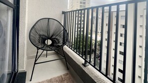 Un balcón con una silla de metal negro, una pared blanca con textura y vistas a edificios al fondo.