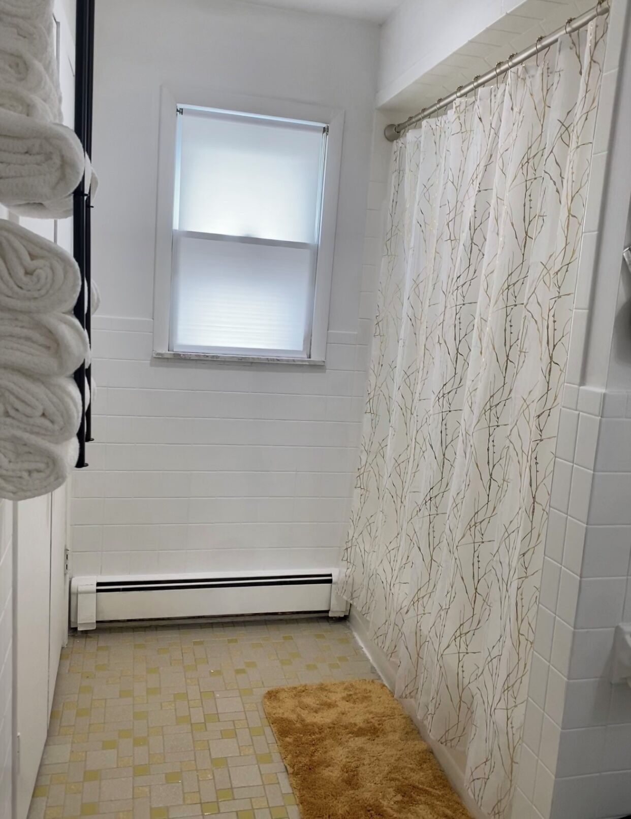Un baño con ventana, cortina de ducha blanca con diseño de ramas, toallero con toallas blancas y una alfombra marrón en el suelo.