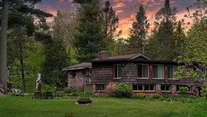 Una casa de madera rústica rodeada de un paisaje verde exuberante y altos árboles, con un cielo al atardecer de fondo.