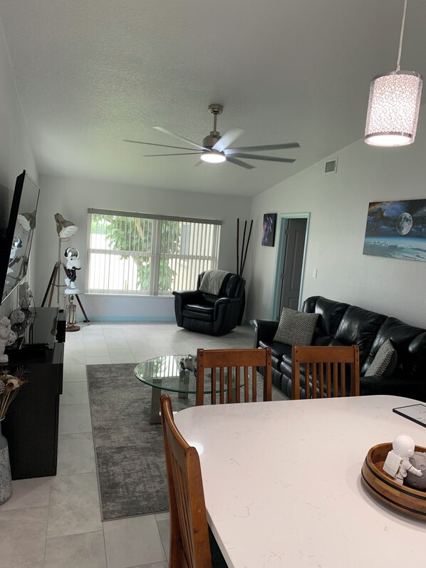 A modern living room with a glass dining table, a black leather sofa, a ceiling fan, and a large window with blinds. - 321 lift off, rockets and dolphins from your window (Cocoa Beach)