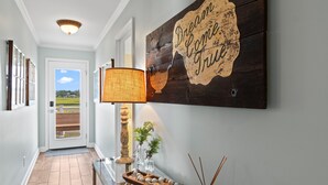 Un corridoio con un cartello di legno che recita “Dream Come True”, un tavolo in vetro con oggetti decorativi e vista su una zona esterna verde attraverso una porta.