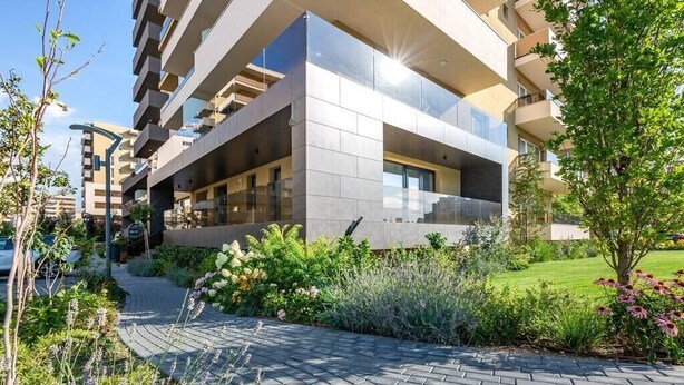 Un immeuble résidentiel moderne à plusieurs étages, doté de balcons, d’une allée pavée et d’une végétation luxuriante.