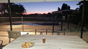 A dining table with a plate of food and a glass of beer, overlooking a swimming pool and a fenced area with trees and a building in the background.