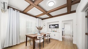 Een eethoek met een houten tafel en stoelen, witte gordijnen en een keuken op de achtergrond.