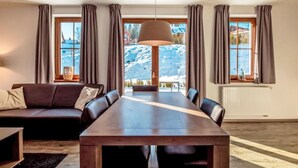 A modern living room with a wooden dining table, leather sofa, and large windows with curtains. - Residence Lipno by Interhome (Lipno nad Vltavou)