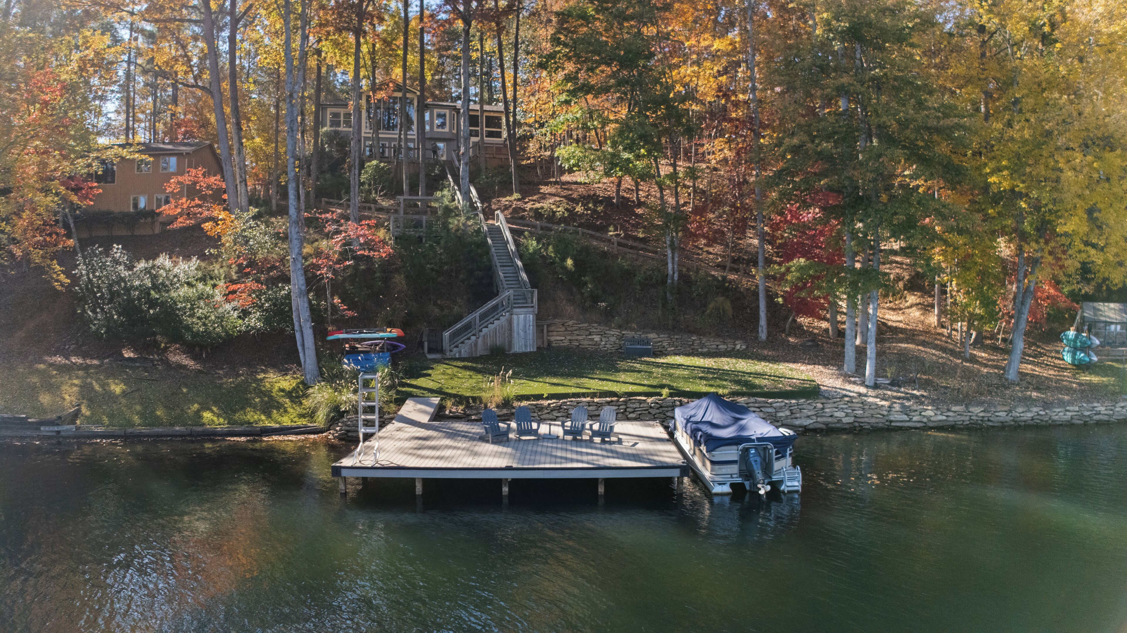 A dock with a covered boat, a stone wall, and a grassy area with Adirondack chairs.
