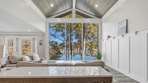 Ein modernes Wohnzimmer mit großem Fenster, Holzbalkendecke und Blick auf Bäume und See.