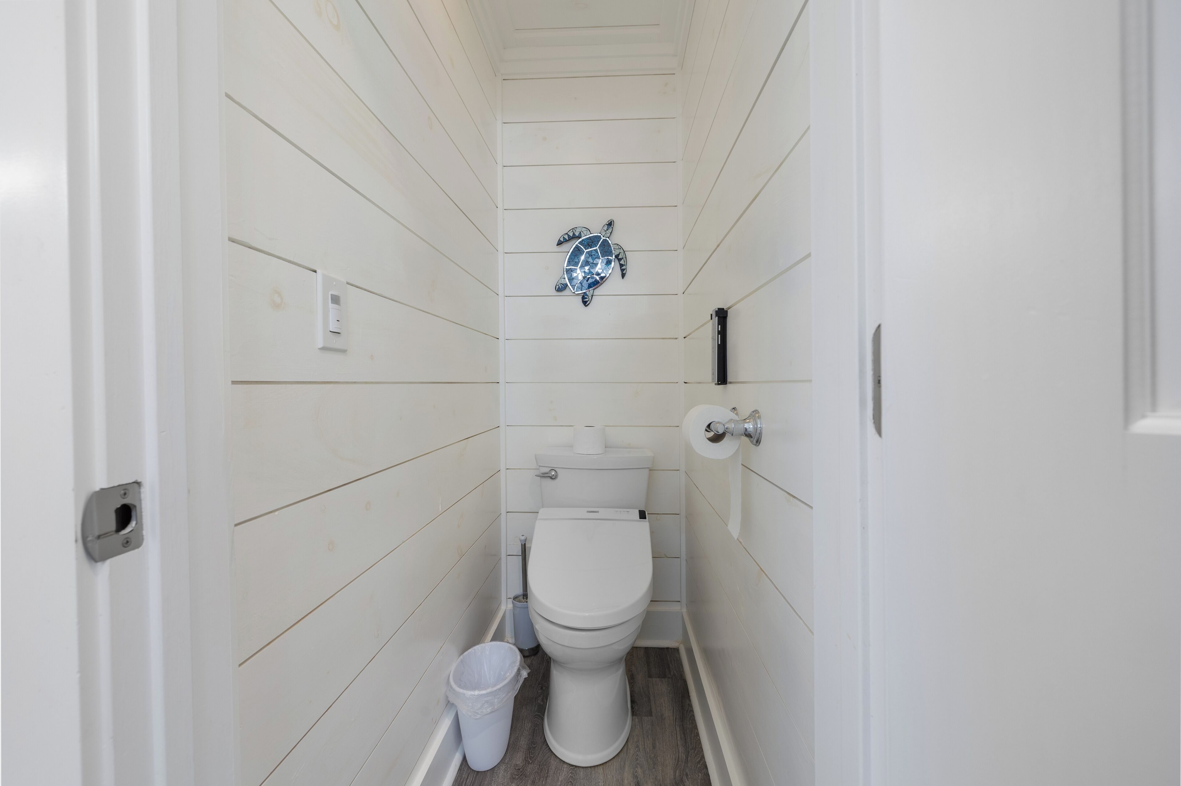 A small, narrow bathroom with white wooden walls, a white toilet, a trash bin, and a wall-mounted toilet paper holder.