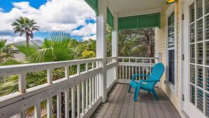 Un porche cubierto con una silla Adirondack azul, barandillas blancas y vistas a palmeras y una casa.