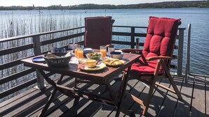 A dining table with breakfast items and a red chair on a wooden deck overlooking a lake. - Bootshaus auf dem See by Interhome (Lindow)