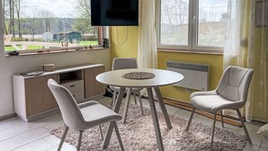 A room with a TV mounted on the wall, a dining table with chairs, a small cabinet, and a view of the outdoors. - am Pälitzsee by Interhome (Rheinsberg)