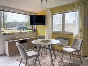 A room with a TV mounted on the wall, a dining table with chairs, a small cabinet, and a view of the outdoors.