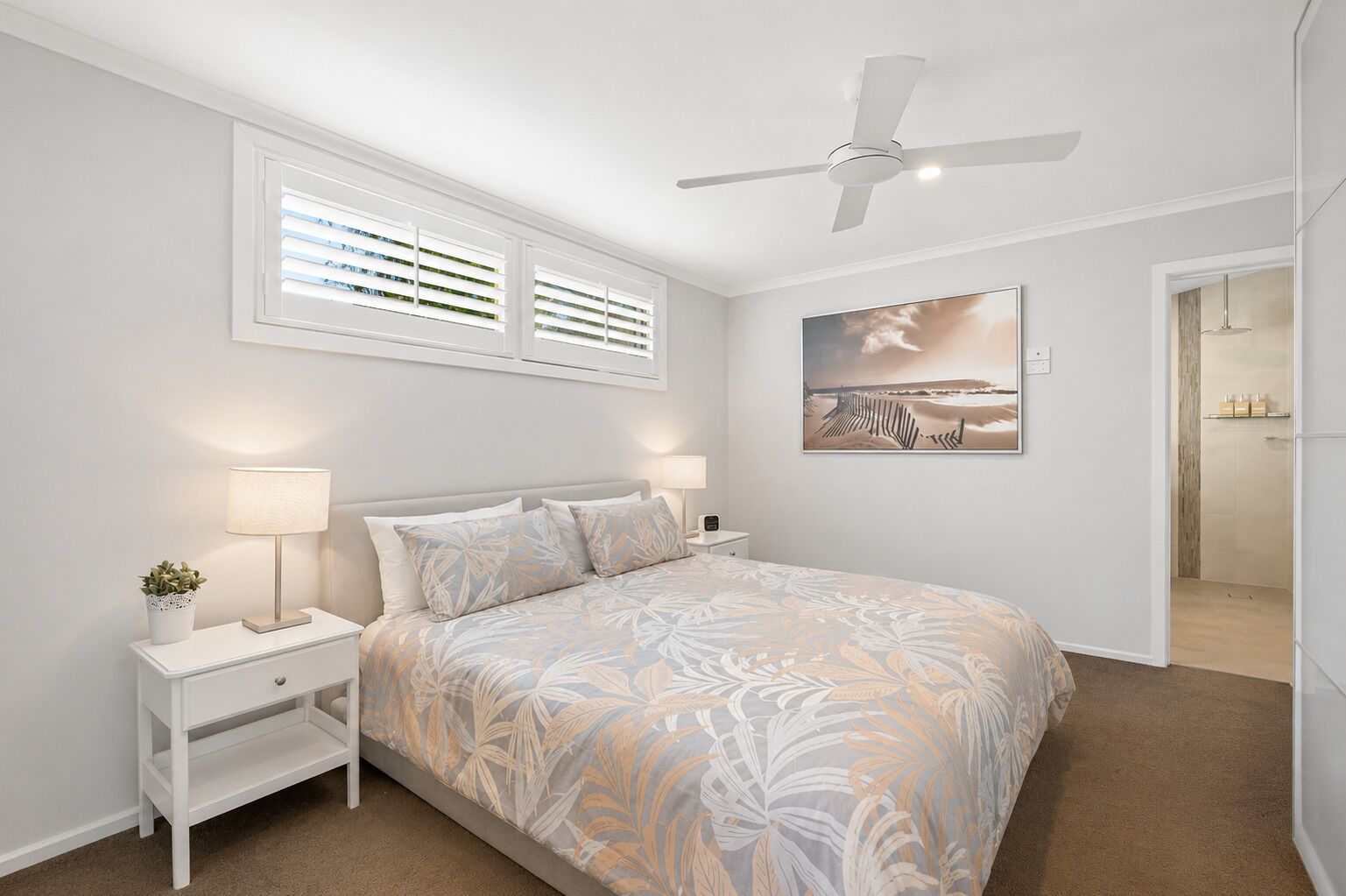 A bedroom with a bed, bedside tables, a ceiling fan, a window with blinds, a painting, and a bathroom visible through an open door.
