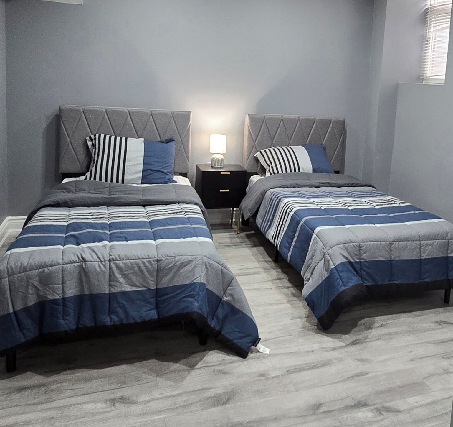 A twin bedroom with two beds, each with a striped duvet and pillows, a nightstand with a lamp, and a window with blinds.
